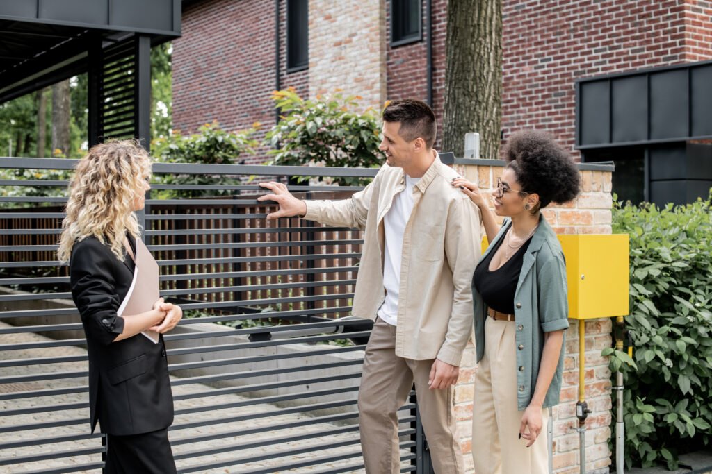 pleased multiethnic couple standing near real estate agent and fence of new city cottage